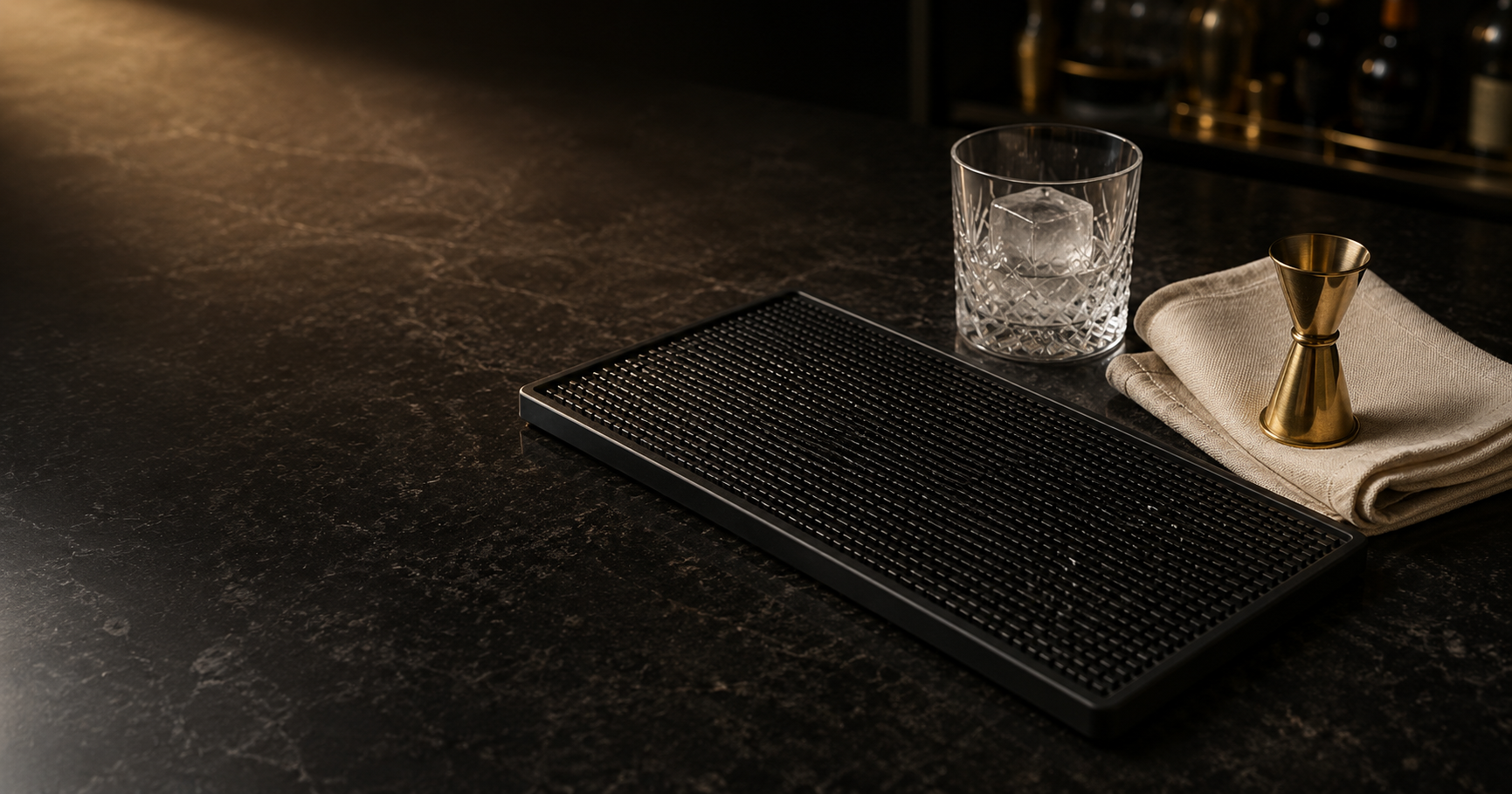 Black textured rubber bar mat on a dark stone counter beside a crystal rocks glass, brass jigger and folded linen towel